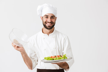 Wall Mural - Image of joyful chief man in cook uniform smiling and holding plate with vegetable salad isolated over white wall