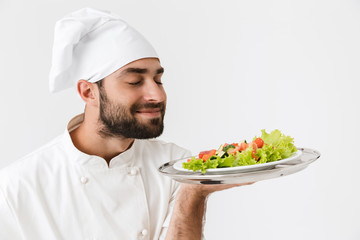 Wall Mural - Image of pleased chief man in cook uniform smelling dish while holding plate with vegetable salad