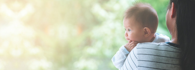 Young asian mother carrying her male toddler with green trees and garden in background. Toddler looking to the left with hand in mouth. Banner style frame with copy space.