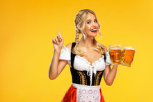 Young Sexy Oktoberfest Girl Waitress, Wearing A Traditional Bavarian Or German Dirndl, Serving Big Beer Mugs With Drink Isolated On Yellow Background. Woman Pointing To Looking Up.
