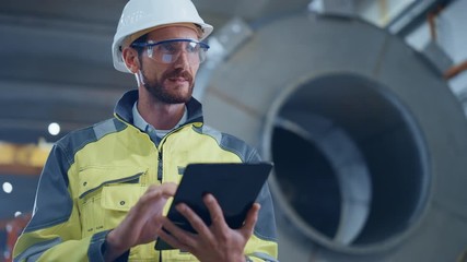 Wall Mural - Portrait of Professional Heavy Industry Engineer / Worker Wearing Safety Uniform and Hard Hat Uses Tablet Computer. In the Background Construction Factory for Oil, Gas and Fuels Transport Pipeline