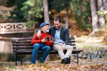 Wall Mural - Senior father and his son sitting on bench by lake in nature, using tablet.