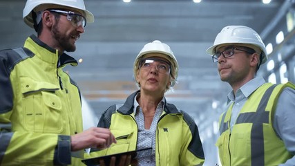 Wall Mural - Three Heavy Industry Engineers Stand in Pipe Manufacturing Factory, Use Digital Tablet Computer, Have Discussion. Large Pipe Assembled. Design and Construction of Oil, Gas and Fuels Transport Pipeline