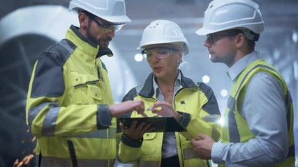 Wall Mural - Three Heavy Industry Engineers Stand in Pipe Manufacturing Factory, Use Digital Tablet Computer, Have Discussion. Large Pipe Assembled. Design and Construction of Oil, Gas and Fuels Transport Pipeline