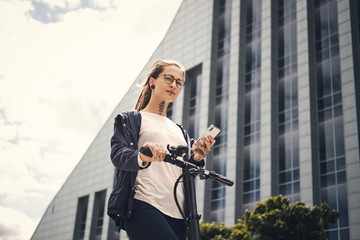 Wall Mural - Attractive pensive girl is chatting by mobile phone while driving electro scooter near interesting building.