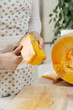Kobiece dłonie trzymają kawałek dyni i obierają go ze skóry za pomocą noża kuchennego. Połowa świeżej dużej pomarańczowej dyni leży na kuchennym blacie.