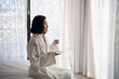 © romankosolapov - Attractive young girl in bathrobe holding a cup, looking at window and smiling while sitting on a bed near the big window at home