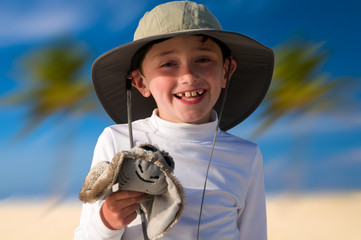 Young smiling young boy at the beach holding a stuffed animal with palm trees in the background.