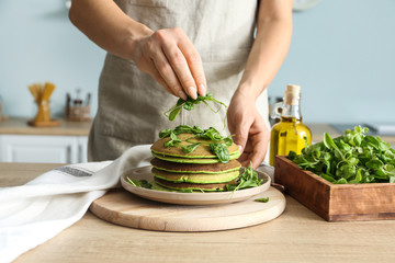 Woman adding spinach to tasty green pancakes in kitchen
