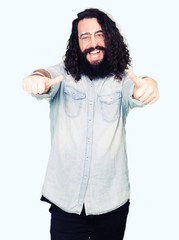 Young hipster man with long hair and beard wearing glasses approving doing positive gesture with hand, thumbs up smiling and happy for success. Looking at the camera, winner gesture.