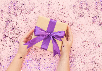 Wall Mural - Child's hands holding giftbox tied with purple color ribbon on pink background with glitter. Flat lay style.