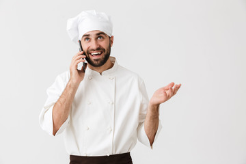 Wall Mural - Emotional happy young chef posing isolated over white wall background in uniform talking by mobile phone.