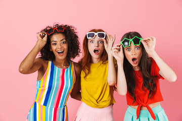 Sticker - Shocked surprised amazing young three multiethnic girls friends posing isolated over pink wall background.