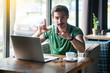 You are loser. Young serious businessman in green t-shirt sitting, pointing and showing loser gesture and looking at camera. business and freelancing concept. indoor shot near big window at daytime.