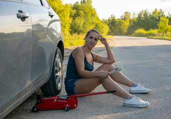 Wall Mural - A beautiful young girl tries to Jack the car to replace the wheel on the road to continue her journey.