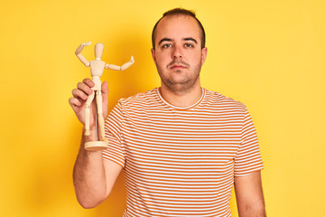 Poster - Young man holding figure of art dummy standing over isolated yellow background with a confident expression on smart face thinking serious