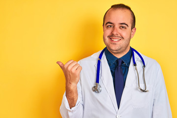 Young doctor man wearing coat and stethoscope standing over isolated yellow background smiling with happy face looking and pointing to the side with thumb up.