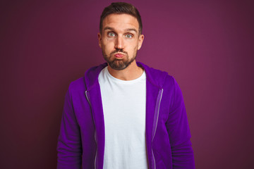 Wall Mural - Young fitness man wearing casual sports sweatshirt over purple isolated background puffing cheeks with funny face. Mouth inflated with air, crazy expression.