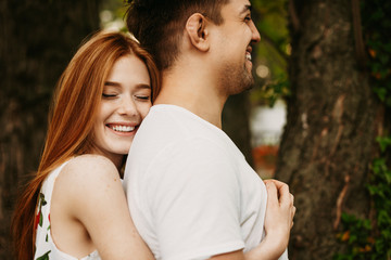 Wall Mural - Amazing red hair woman with freckles laughing with closed eyes while embracing her boyfriend from back which is laughing outdoor in the city.