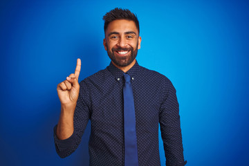 Wall Mural - Young indian businessman wearing elegant shirt and tie standing over isolated blue background showing and pointing up with finger number one while smiling confident and happy.