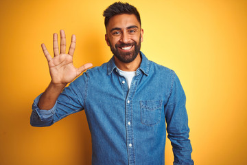 Wall Mural - Young indian man wearing denim shirt standing over isolated yellow background showing and pointing up with fingers number five while smiling confident and happy.