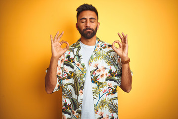 Young indian man on vacation wearing summer floral shirt over isolated yellow background relax and smiling with eyes closed doing meditation gesture with fingers. Yoga concept.