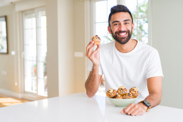 Wall Mural - Handsome hispanic man eating chocolate chips muffin with a happy face standing and smiling with a confident smile showing teeth