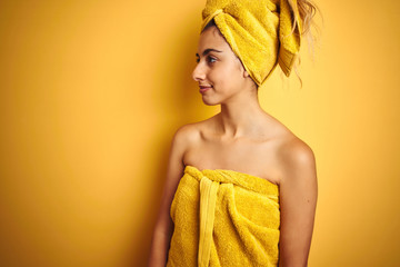 Poster - Young beautiful woman wearing a shower towel after bath over yellow isolated background looking to side, relax profile pose with natural face with confident smile.