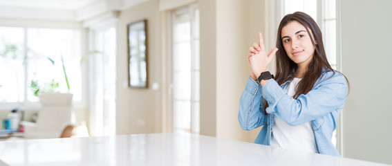 Poster - Wide angle picture of beautiful young woman sitting on white table at home Holding symbolic gun with hand gesture, playing killing shooting weapons, angry face