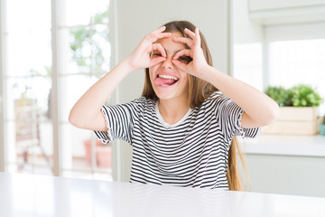 Wall Mural - Beautiful young girl kid wearing stripes t-shirt doing ok gesture like binoculars sticking tongue out, eyes looking through fingers. Crazy expression.