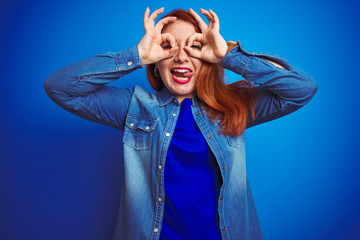 Sticker - Young beautiful redhead woman wearing denim shirt standing over blue isolated background doing ok gesture like binoculars sticking tongue out, eyes looking through fingers. Crazy expression.