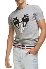 Cropped medium full shot of a young man dressed in a gray graphic tee with painted deers and a big snowflake and light blue jeans with a belt. The textile belt with blue, white and red stripes