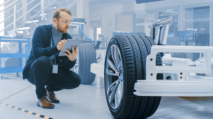 Leinwandbilder - Engineer with Glasses and Beard Works on a Tablet Computer Next to an Electric Car Chassis Prototype with Wheels, Batteries and Engine in a High Tech Development Laboratory.