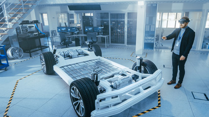 Fototapete - Automotive Engineer Working on Electric Car Chassis Platform, Using Augmented Reality Headset. In Innovation Laboratory Facility Concept Vehicle Frame Includes Wheels, Suspension, Engine and Battery.