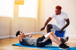 © twinsterphoto - Side view of Asian man lying on floor doing abdominal exercises while African American trainer kneeling besides and teaching in fitness club