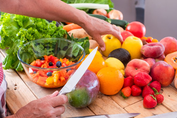 Two hands of a senior woman working and cutting different kind of fruit and vegetables. Bright light frow the window. Wooden table with a large group of colorful fruits and vegetables. Healthy eating