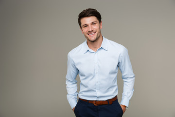 Image of happy brunette man wearing formal clothes smiling at camera with hands in pockets