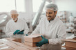 © Dusan Petkovic - Smiling hardworking dedicated Caucasian employee arranging boxes while his superior using tablet and checking on him. Both are dressed in white sterile uniforms. Food plant interior.