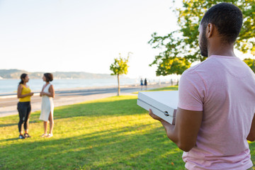 Male courier delivering pizza in park. Back of young man in casual walking on grass to two women and carrying pizza boxes. Pizza order concept