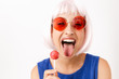Portrait of cute funny woman wearing pink wig and eyeglasses holding lollipop and sticking out her tongue