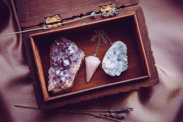 Witch tools inside beautiful old wood box. Rose quartz pendulum, natural amethyst and celestite crystal clusters. Dry lavender flowers on dark purple cloth. Alternative lifestyle concept.