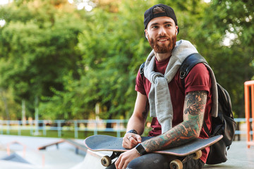 Attractive cheerful young man sitting at the skate park ramp