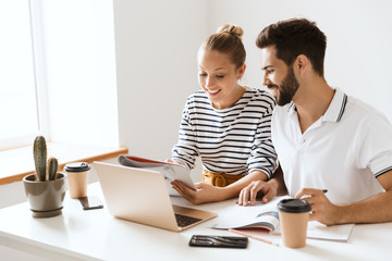 Poster - Optimistic happy pleased young loving couple friends man and woman sitting at the table indoors using laptop computer reading book studying.