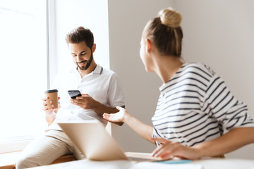 Wall Mural - Blurred woman indoors sitting at the table using laptop computer looking at man using mobile phone on background.