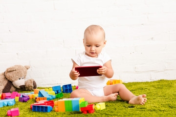 adorbale kid sitting among toys on green floor and holding digital device