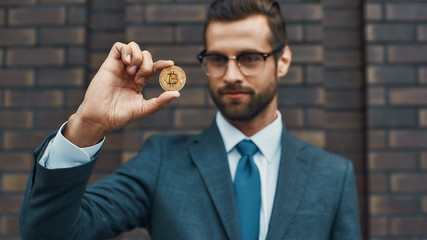 Digital currency. Handsome businessman in formal wear and eyeglasses holding bitcoin in one hand and looking at it while standing against brick wall