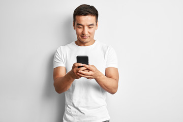 Handsome Asian guy in white studio smiles joyfully looks at the screen of mobile phone. Happy contented man dressed casual writes and reads sms on smartphone