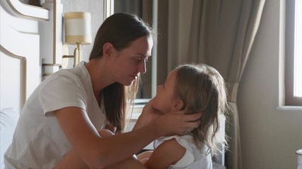 young mother caresses her little daughter in bedroom at home. they sits on the bed together face to 
