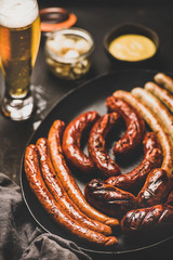 Wall Mural - Octoberfest dinner table concept with grilled veal and pork sausages, sauce in jar, pickled vaggies and beer in glass over dark background, selective focus, close-up