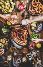 Flat-lay Of Octoberfest Party Dinner Table With Grilled Sausages, Pretzel Pastry, Potatoes, Cucumber Salad, Sauces, Beers And Peoples Hands With Food And Glasses Over Dark Wooden Background, Top View